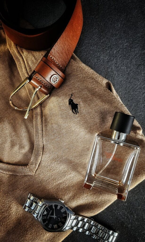 A stylish flatlay showcasing men's accessories: leather belt, wristwatch, and perfume bottle on a beige shirt.